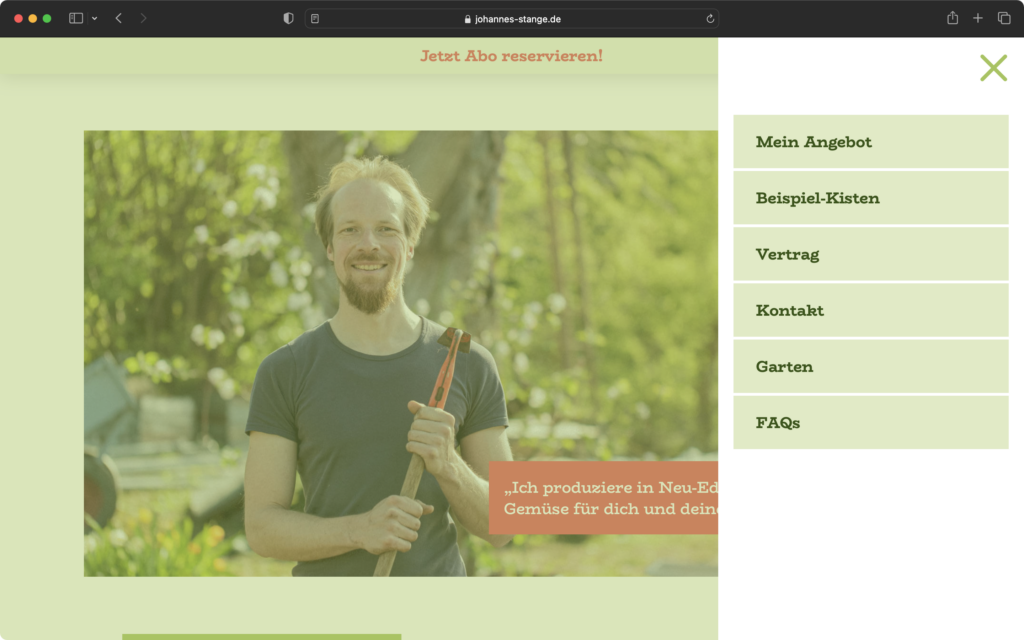 Mann mit Bart und kurzärmeligem Shirt hält Gartengerät und steht lächelnd vor grünem, bewachsenem Hintergrund.