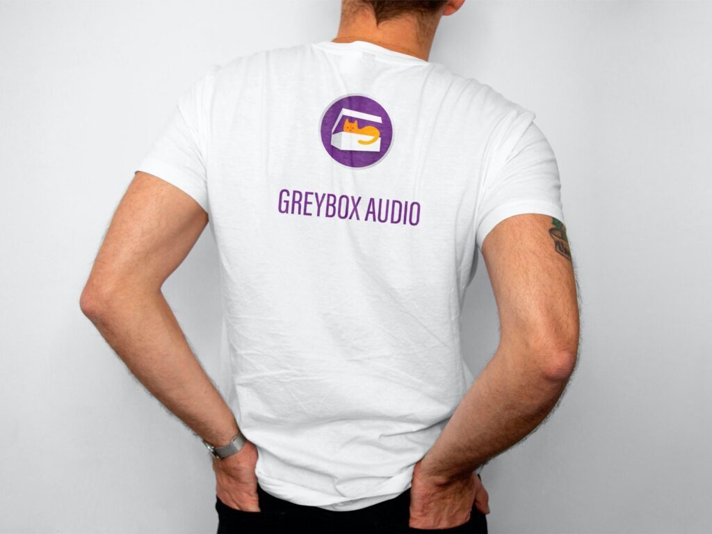 Person von hinten mit weißem T-Shirt, auf dem Rücken ein lila Logo mit einer orangefarbenen Katze in einer Box und der Schriftzug Greybox Audio.