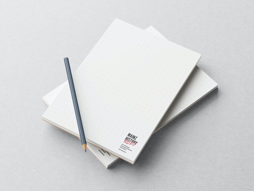 Zwei punktkarierte Notizbücher liegen übereinander auf einem hellen Untergrund, darauf liegt ein grauer Bleistift.