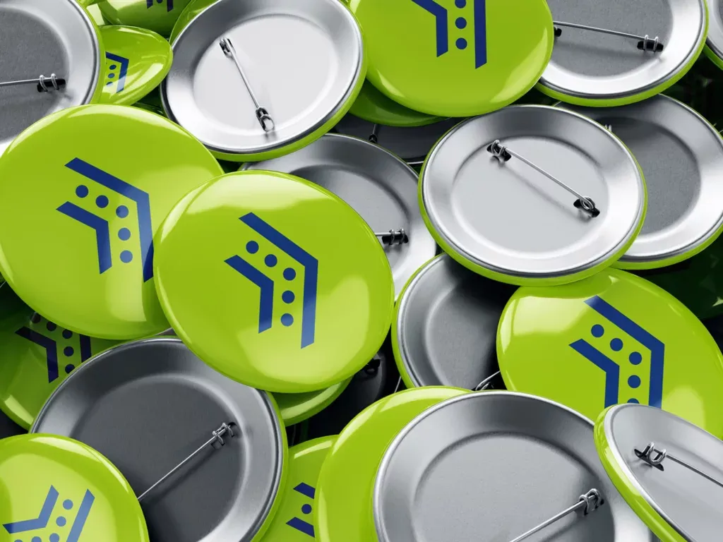 Grüne Ansteckbuttons mit blauem Symbol liegen gemischt mit der Rückseite der Buttons, die Sicherheitsnadeln zeigen.