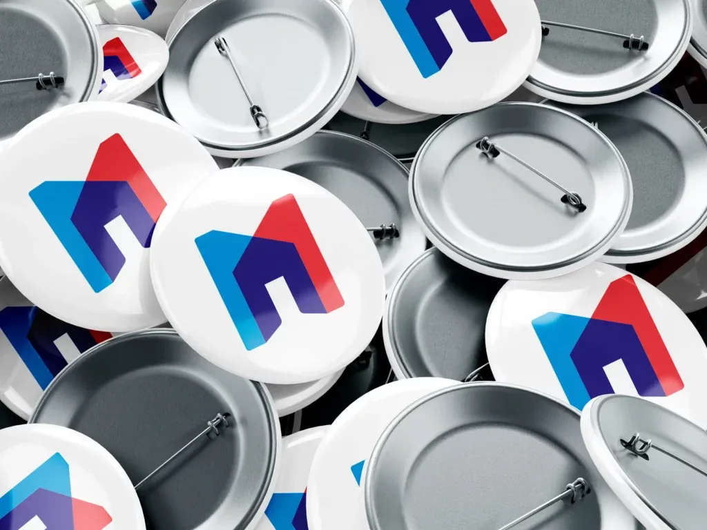 Viele weiße Ansteckbuttons mit einem farbigen Logo in Blau, Rot und Dunkelblau, einige zeigen die Rückseite mit Sicherheitsnadel.