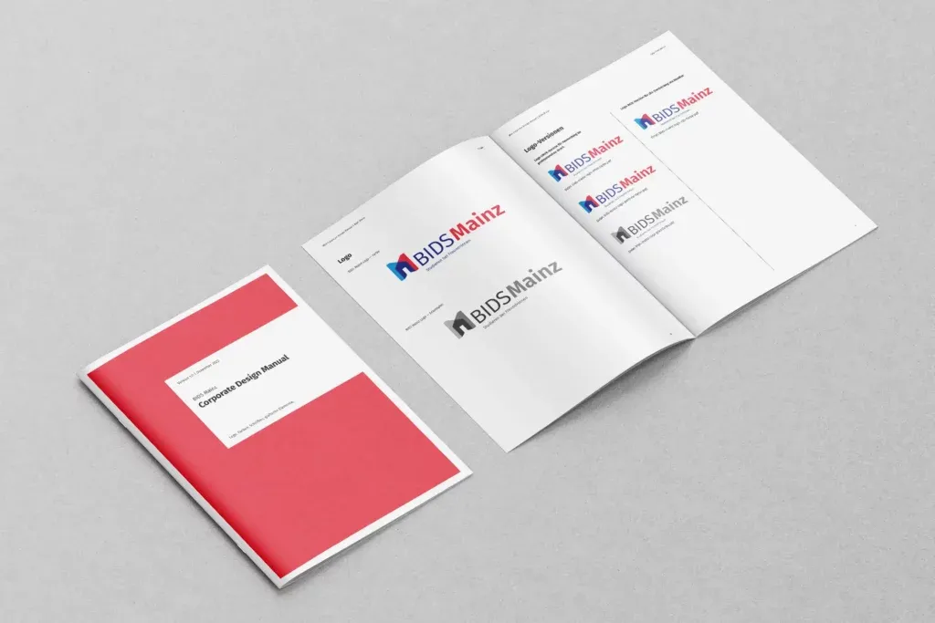 Zwei Corporate Design Manuals von BIDS Mainz liegen auf grauem Untergrund, eines geschlossen mit rotem Cover, das andere geöffnet mit verschiedenen Logo-Versionen.