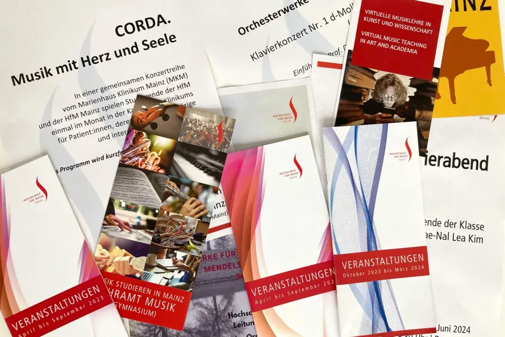 Mehrere Flyer und Broschüren der Hochschule für Musik Mainz mit Veranstaltungshinweisen und Bildern von Musikinstrumenten liegen auf einem Tisch.