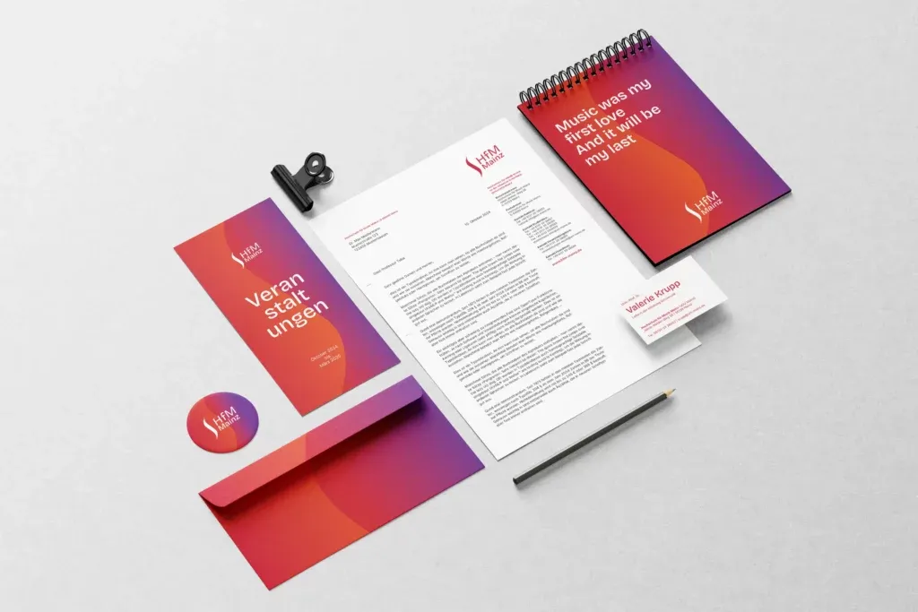 Geschäftsausstattung mit Briefpapier, Visitenkarte, Notizblock, Flyer, Umschlag, Button und Bleistift in Rot- und Violetttönen mit HfM Mainz Logo.